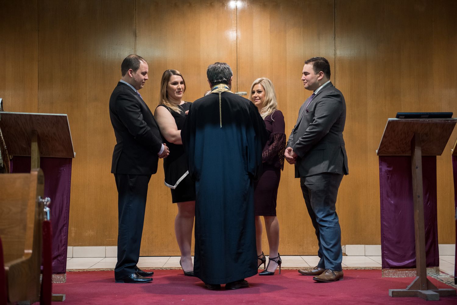 Family gathered around priest in black robe during intimate Orthodox baptism blessing ceremony.