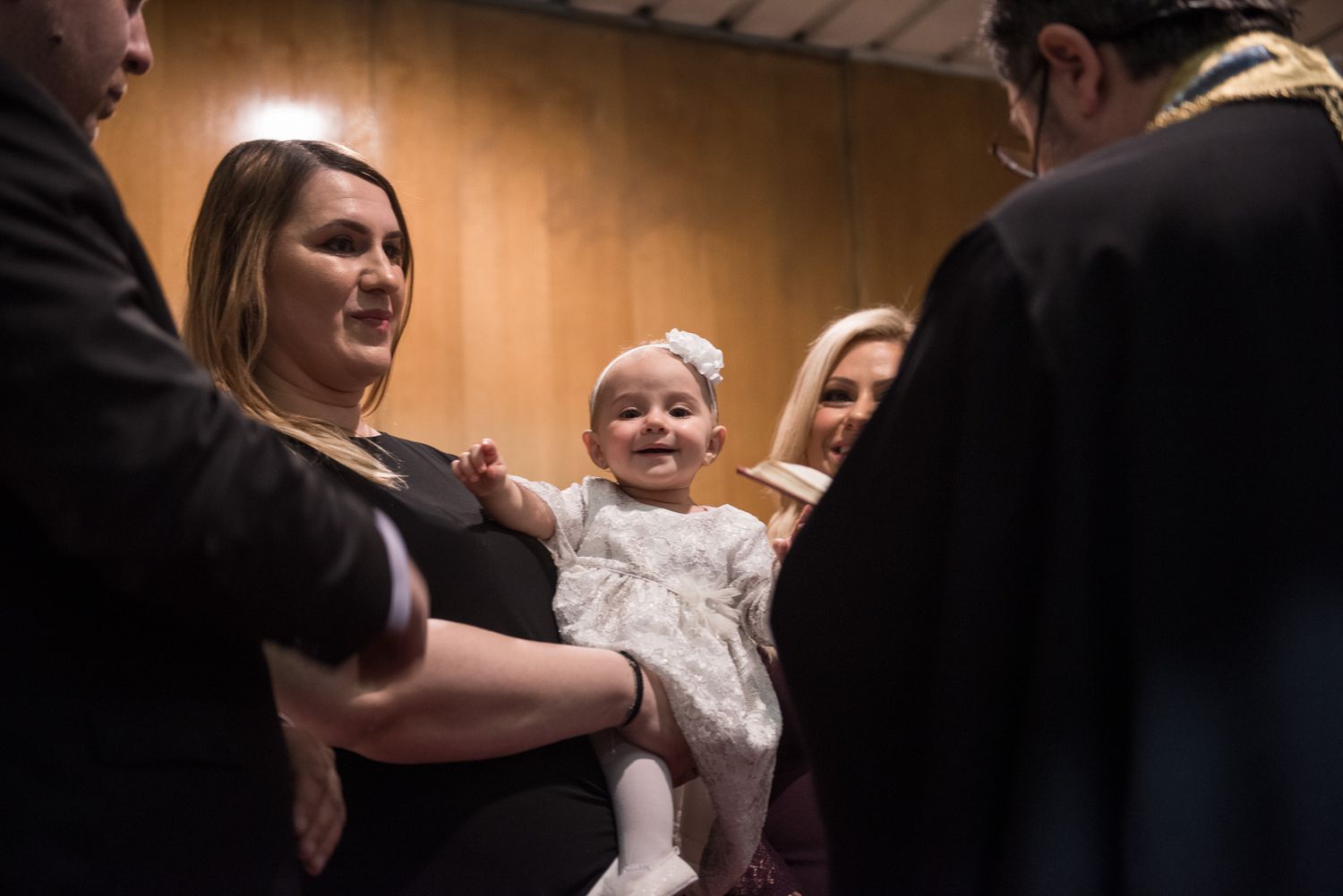 Mother holding smiling baby in white christening gown during baptism ceremony.