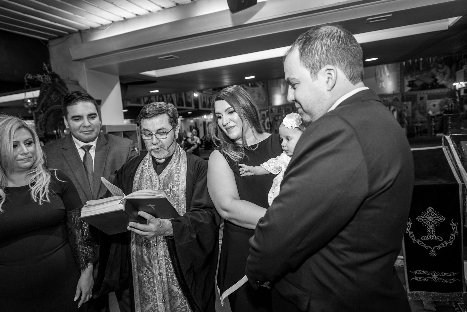 Family gathering around priest reading from book during Greek Orthodox christening reception.