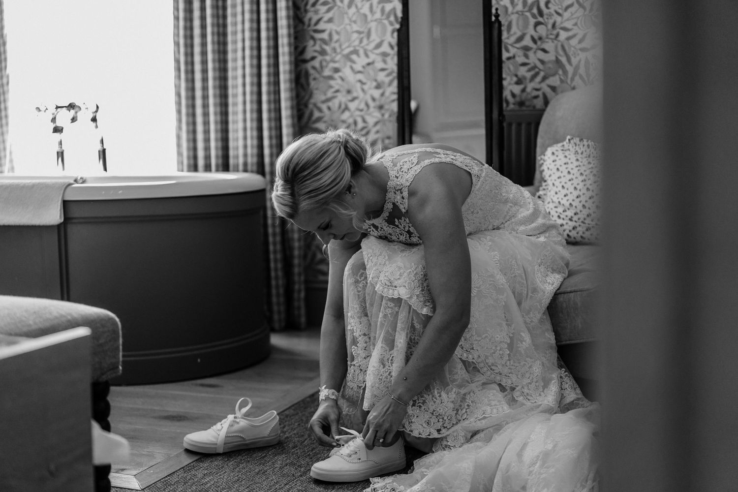Bride in white lace dress tying her wedding shoes in a softly lit room, captured in elegant black and white photography.