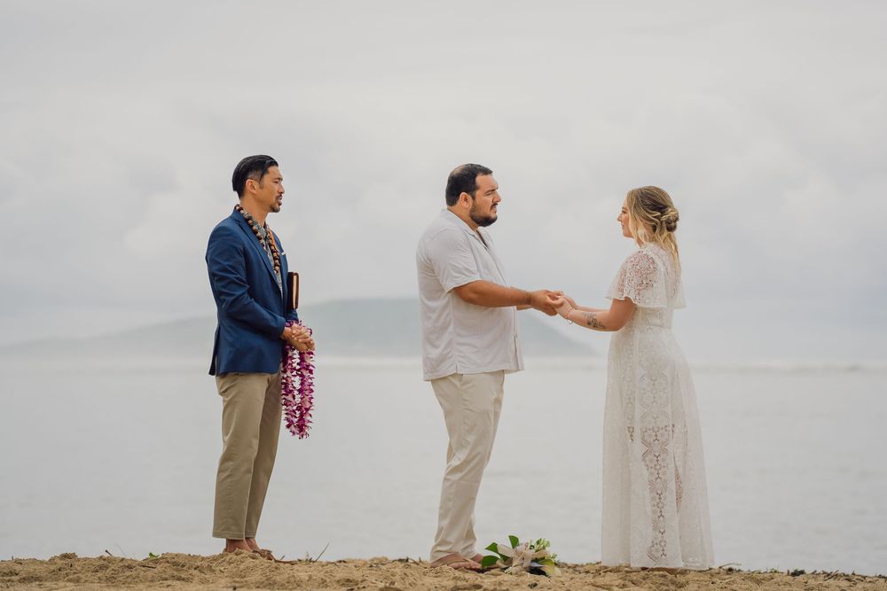Couple exchanging vows in a beach wedding ceremony with an officiant holding a lei, ocean backdrop.