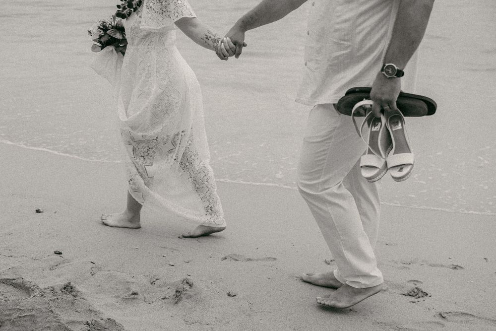 Black and white photo of a bride and groom walking barefoot on a beach, holding hands, groom carrying white heels.