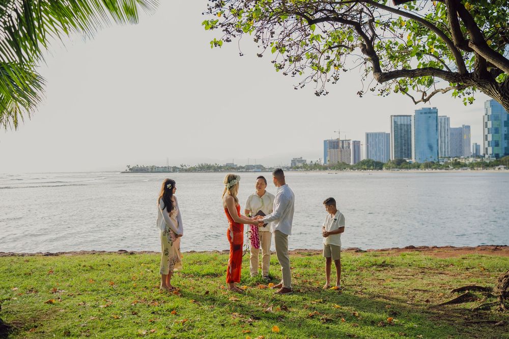 Small wedding ceremony on a grassy waterfront with city skyline, couple exchanging vows under a tree in Hawaii.
