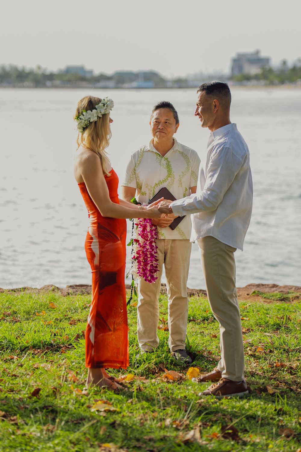 Couple exchanges vows in Hawaiian beach wedding ceremony, bride in orange dress with floral crown, officiant holding book.