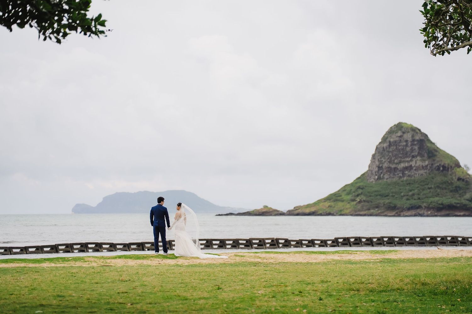 Bride and groom stand by oceanside path with iconic Chinamans Hat island in background on Oahu Hawaii.