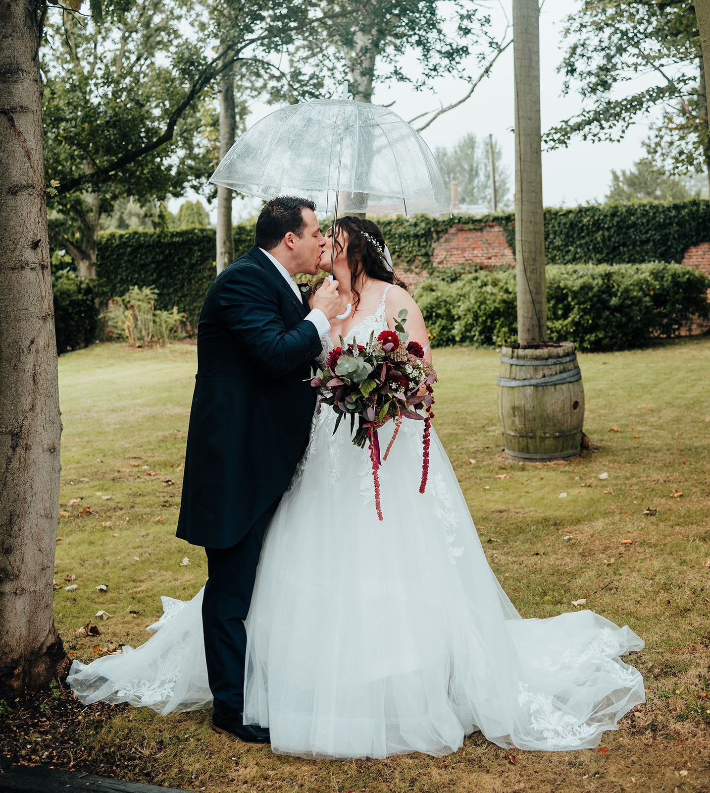 A bride and groom share a kiss under a clear umbrella in a garden, with the bride holding a red and burgundy bouquet.