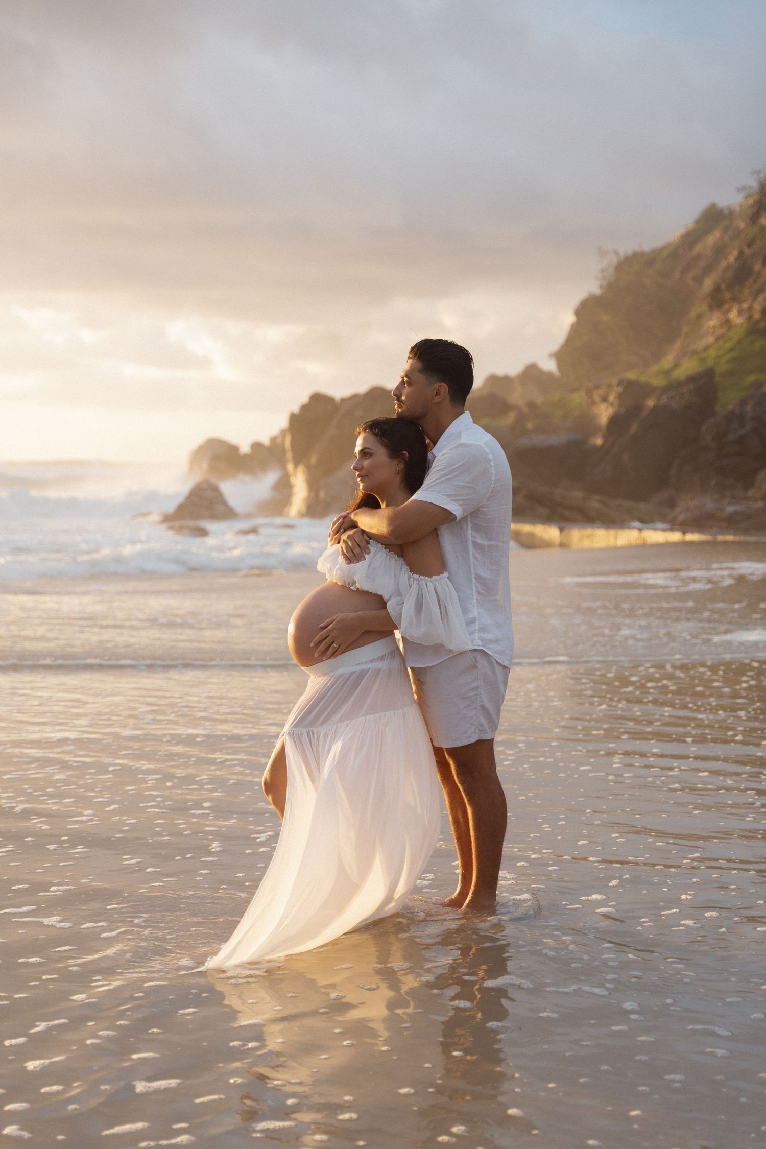 Pregnant couple embracing on a beach at golden sunset, woman in flowing white dress, man in white outfit.