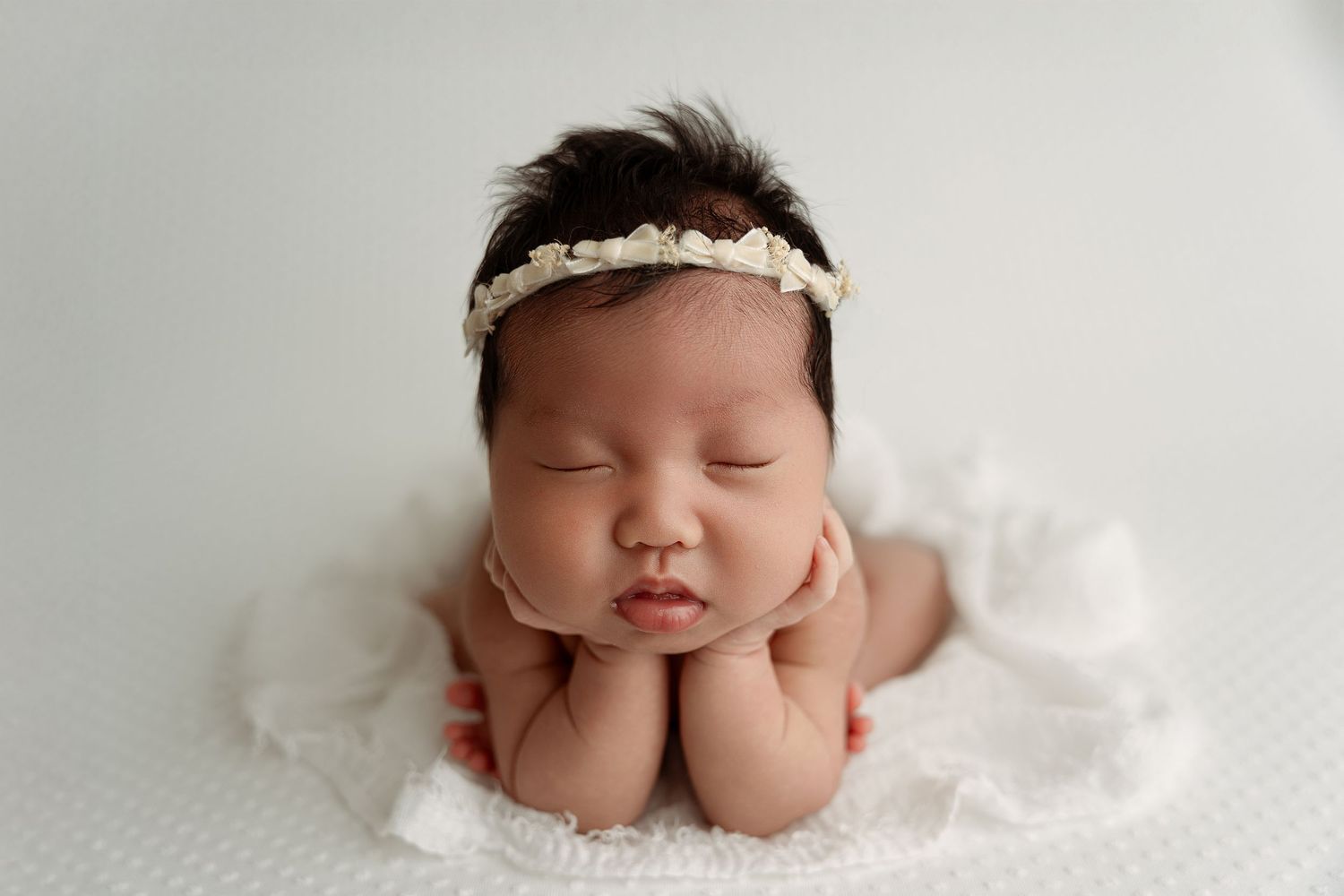 Sleeping newborn baby girl wearing a floral headband, posed with hands under chin on a white backdrop.