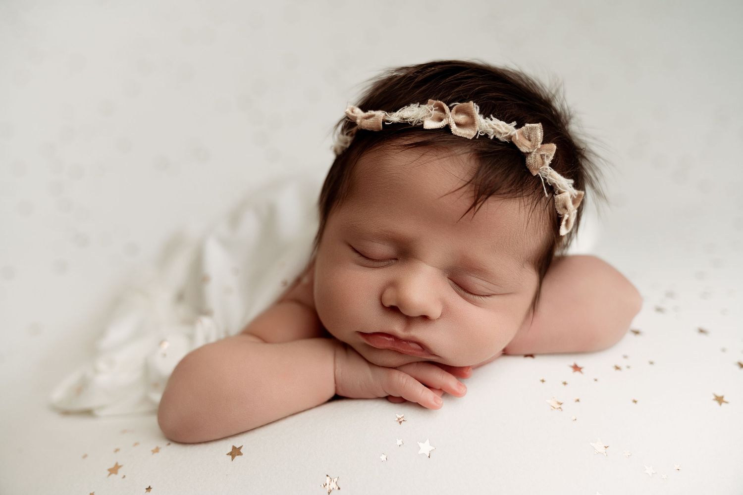 Peaceful newborn baby girl sleeping with chin on hands, wearing a delicate floral headband on a white background.