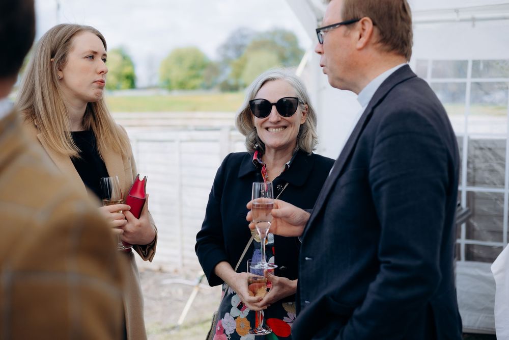 Three elegantly dressed guests socializing outdoors, one holding champagne, at a christening party.