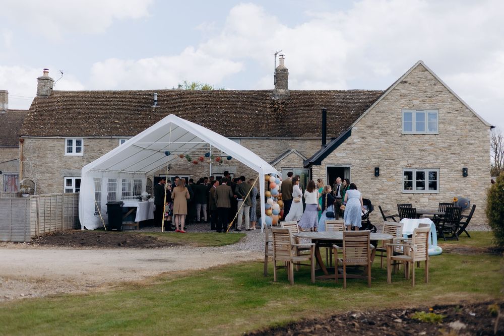 Rustic stone farmhouse wedding venue with white marquee tent and guests gathered outside on a cloudy day.