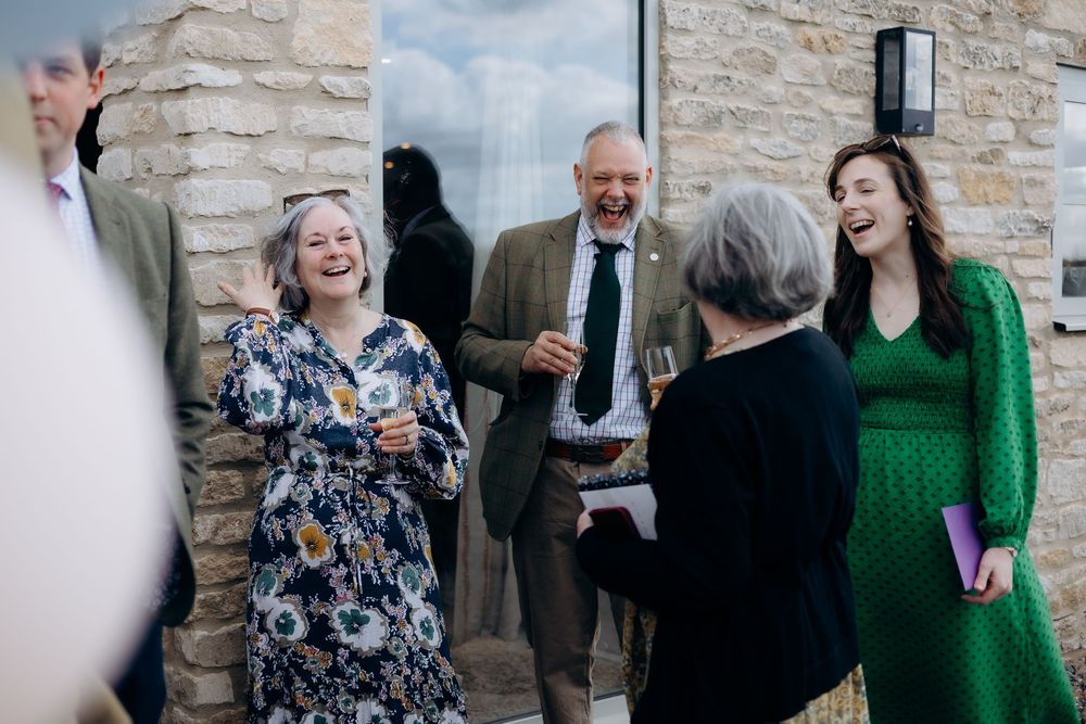 Guests laugh together outside stone building at christening celebration, woman in green dress smiles.