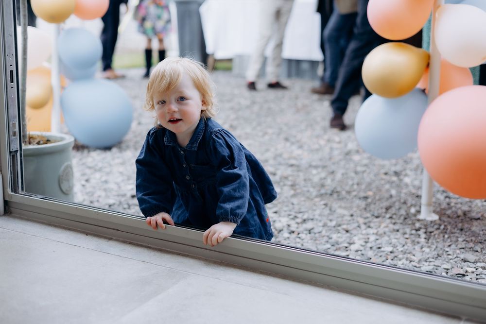 A toddler in a denim outfit peers through a glass door surrounded by colorful balloons.