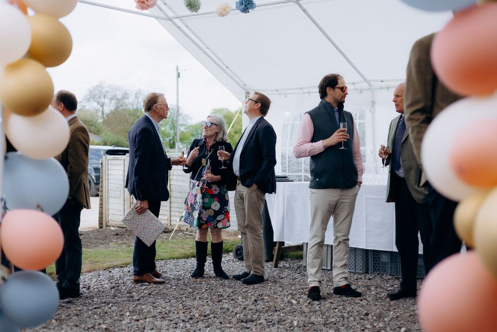 Adults mingling under a marquee tent decorated with pastel balloons at an outdoor event.