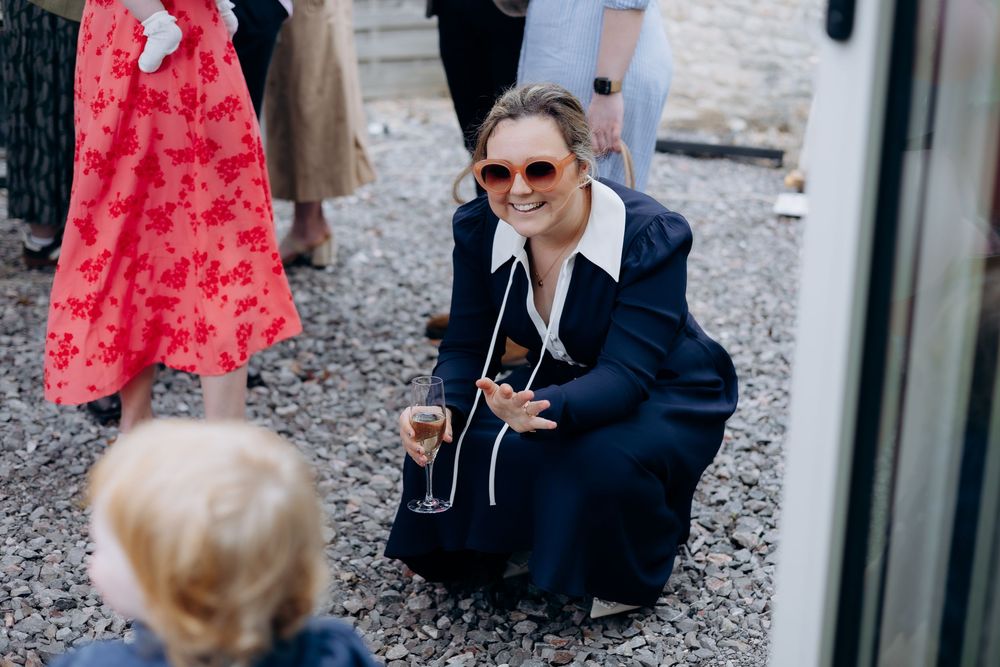 Woman in navy blazer crouching down smiling at child while holding champagne glass at outdoor wedding.