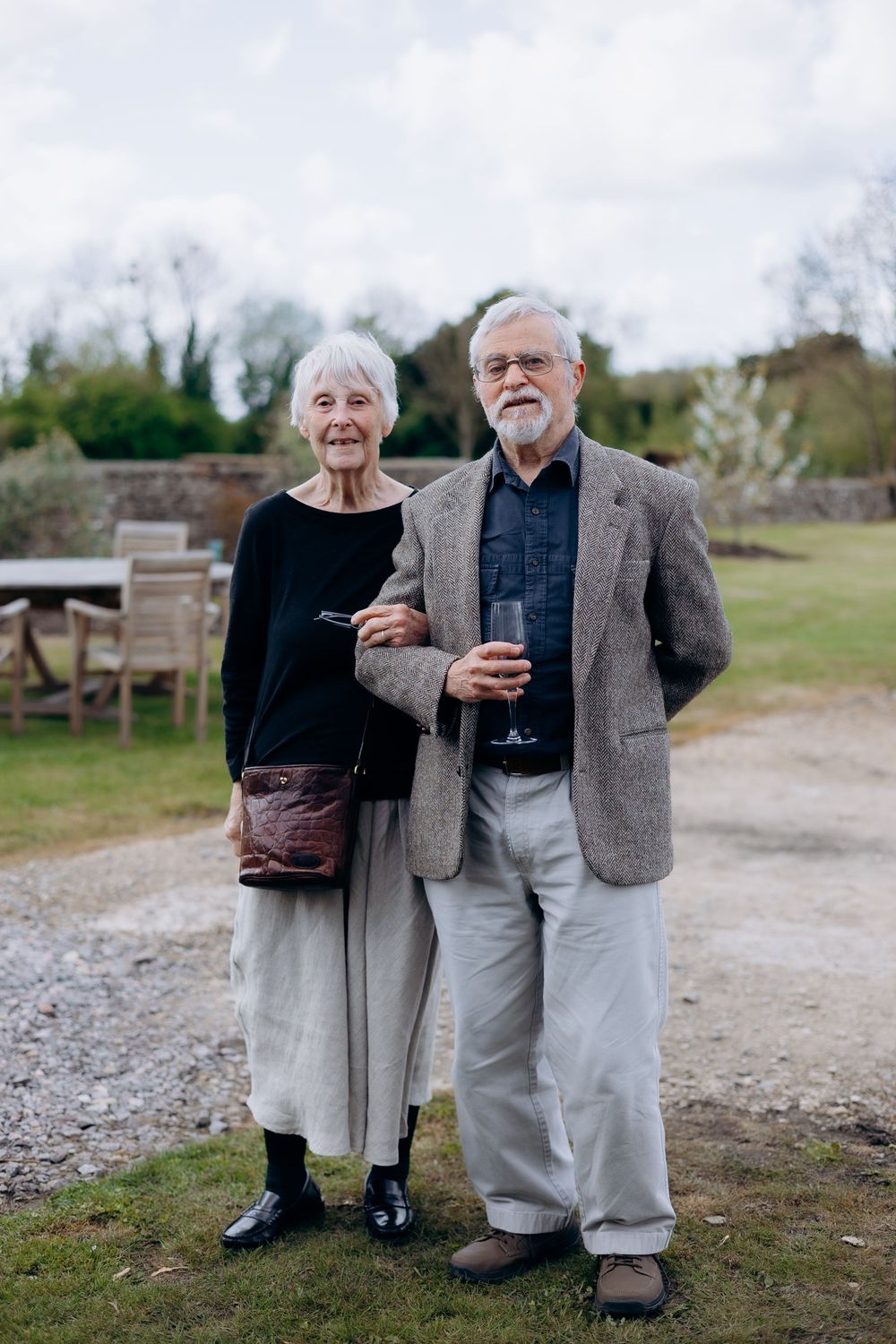 Elegant elderly couple posing together outdoors at a christening celebration in a garden setting.