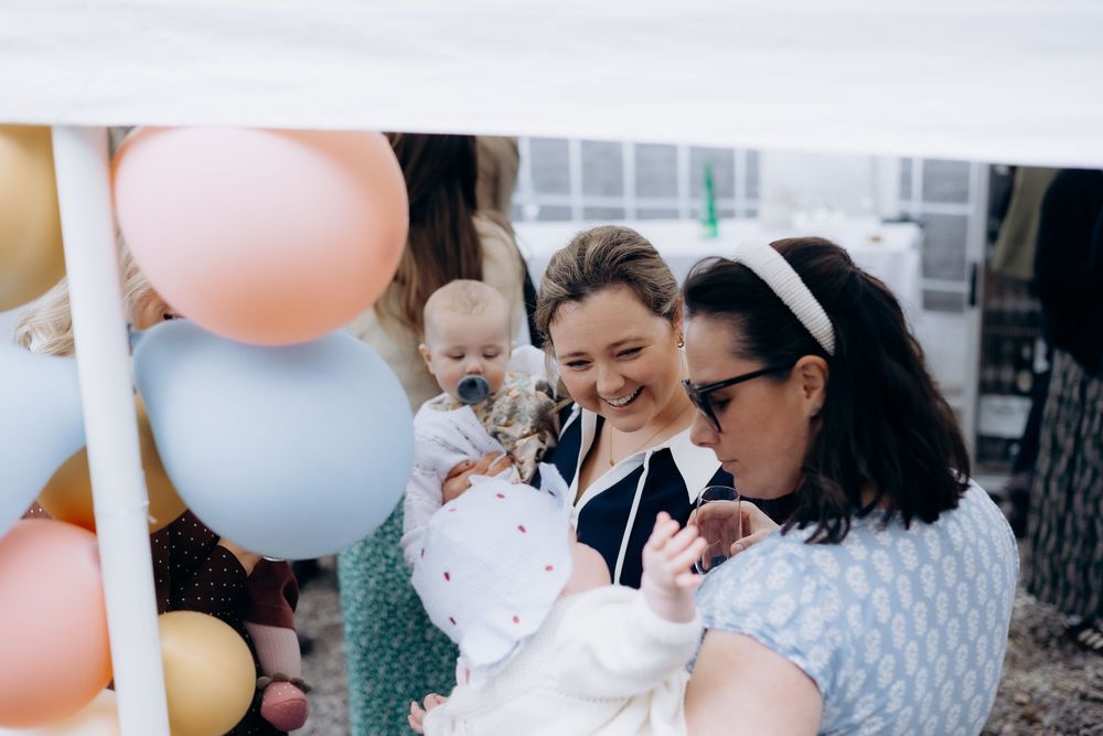 Two women smiling warmly while holding babies under a balloon arch at a colorful celebration event.