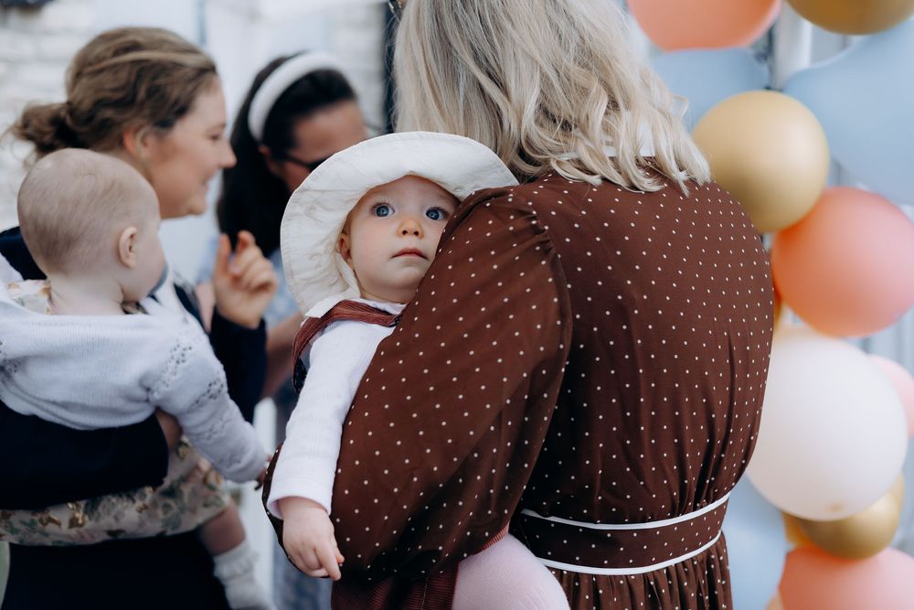 A baby in a white bonnet being carried on an adults back near gold and pink balloons at a party.