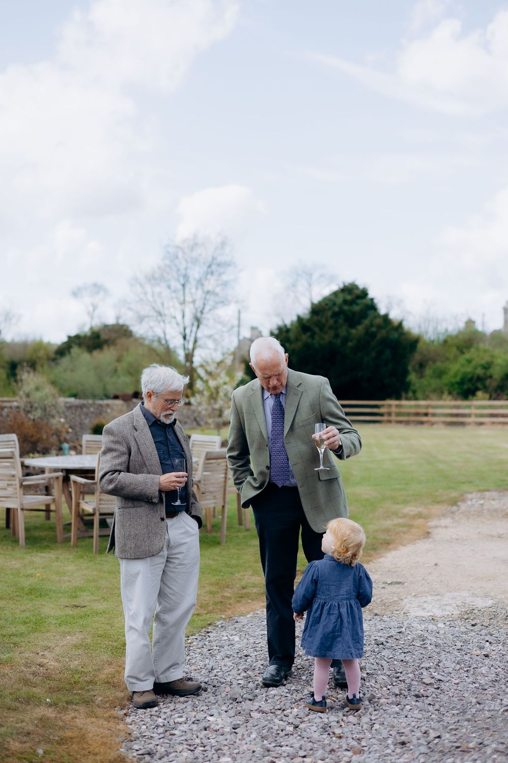 Two elderly gentlemen with drinks chat with a toddler girl in a garden at a christening party.