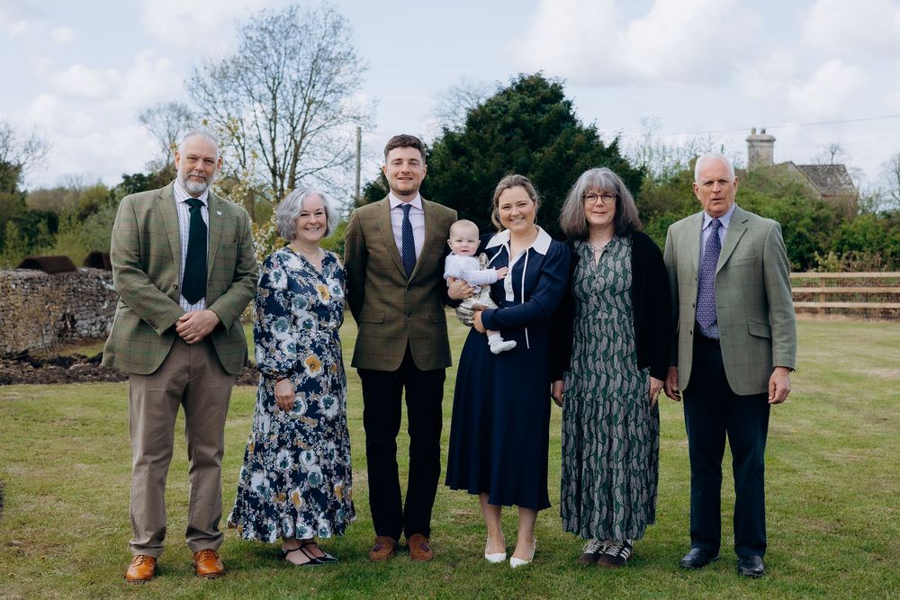 Family of six adults and a baby pose for a formal group portrait outdoors at a christening celebration.