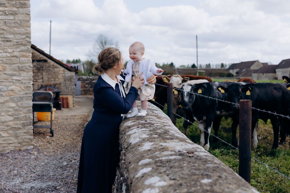 A woman in navy dress holds a baby near stone fence with black and white cows on a farm.