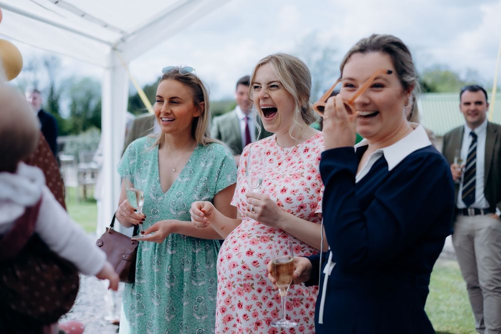 Three women laughing joyfully at an outdoor wedding reception under a marquee tent.