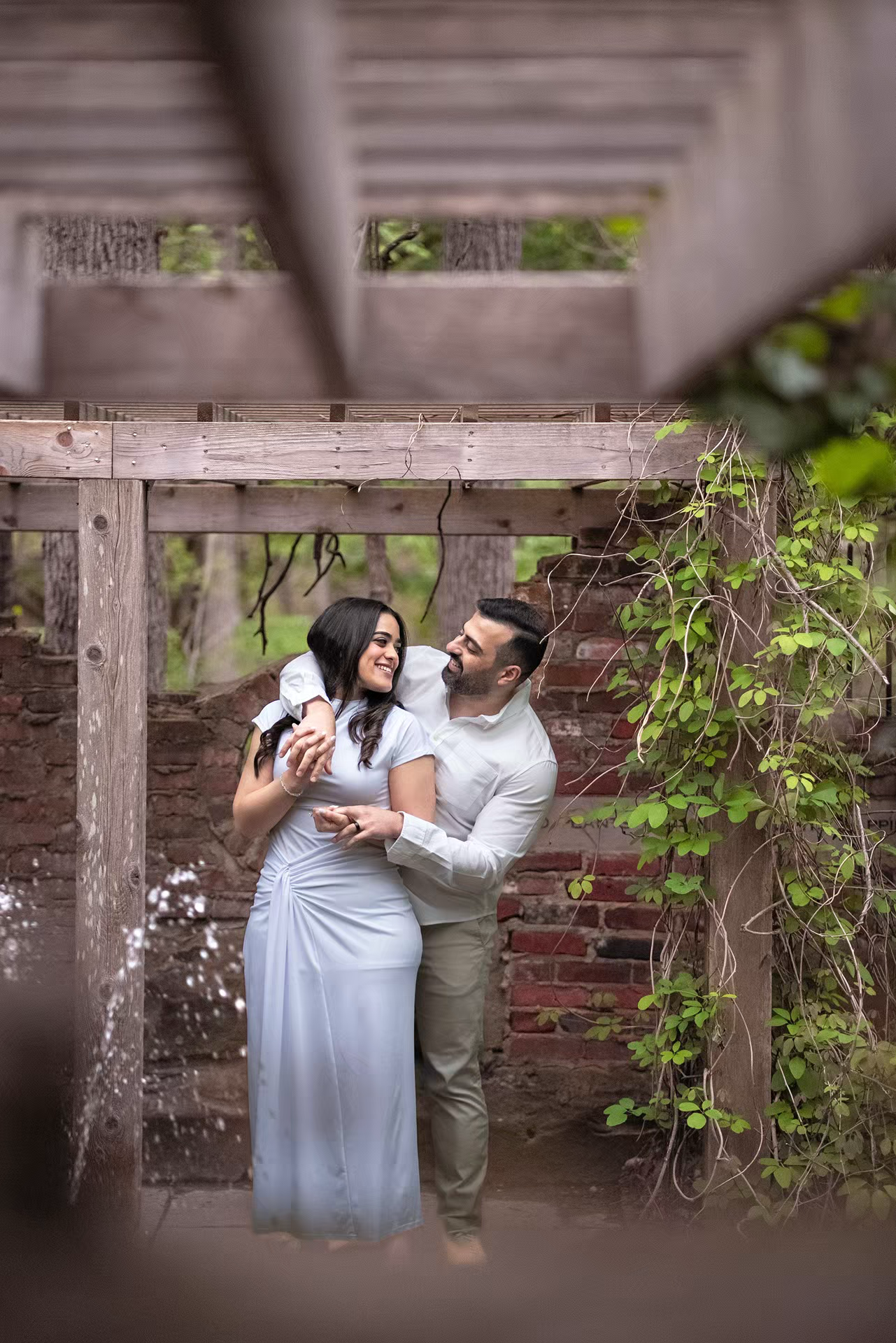 Couple embracing and smiling under a rustic wooden pergola with ivy-covered brick walls in a lush garden setting.
