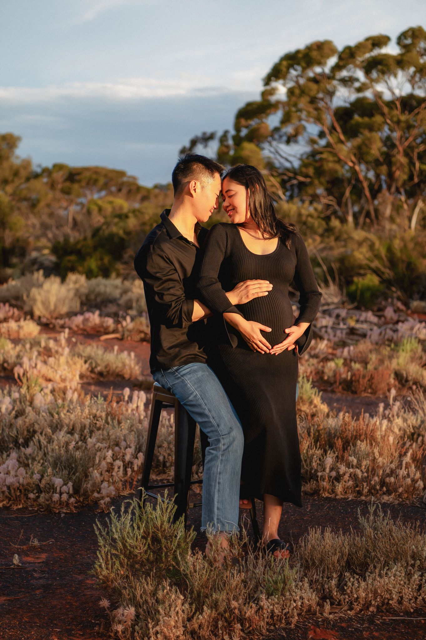 Couple in black outfits sharing a tender moment outdoors, woman cradling pregnant belly at golden hour in Australian bush.