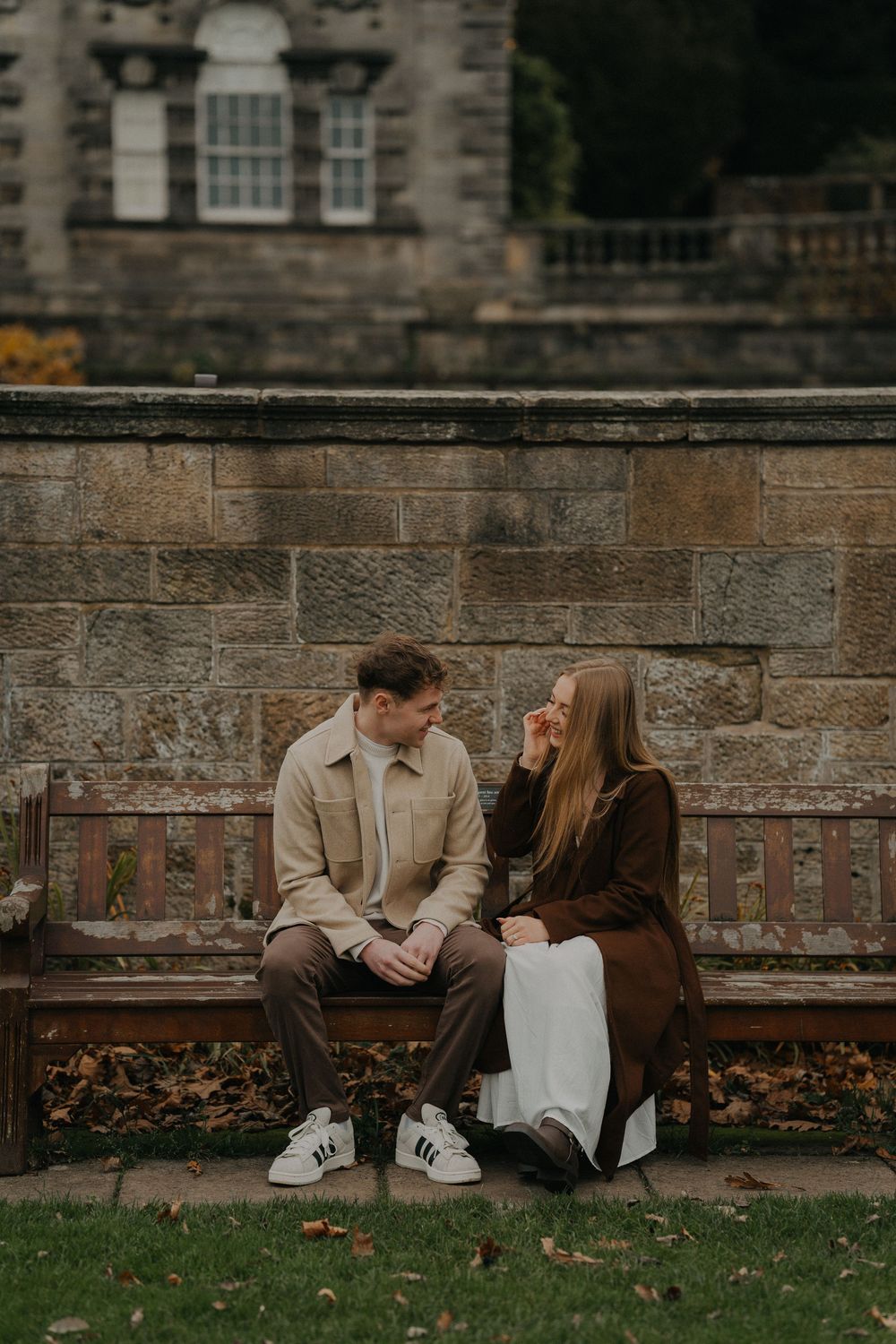 Couple sitting on a wooden bench against a stone wall in an autumn park, sharing an intimate conversation.