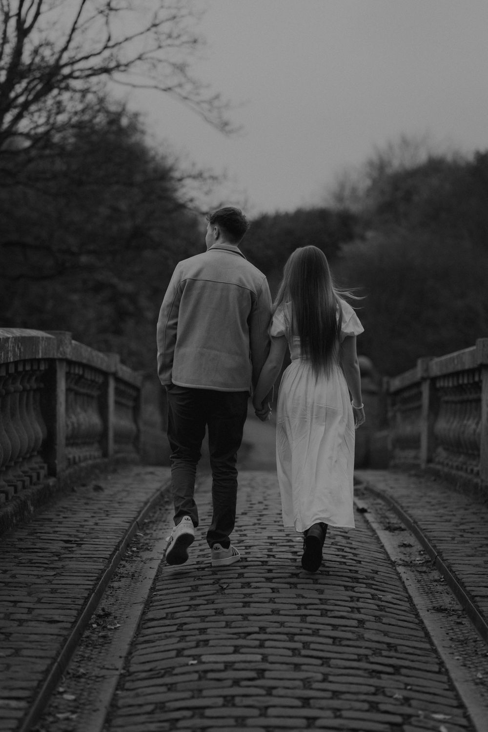 Black and white photo of a couple holding hands walking away on a cobblestone bridge surrounded by autumn trees.