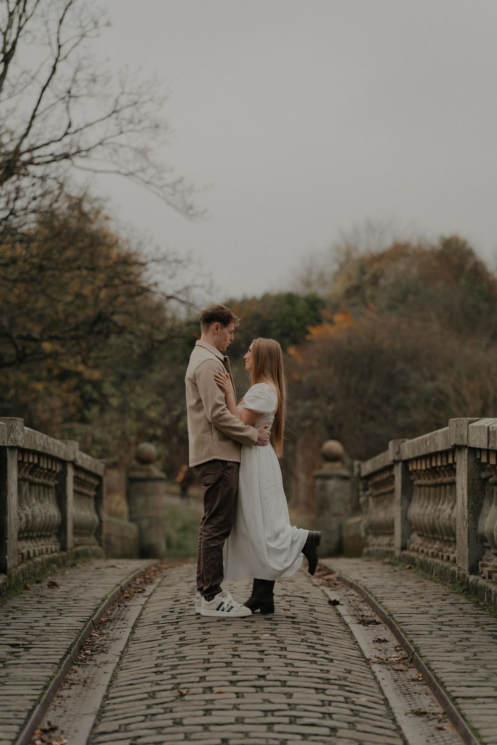 Couple embracing on a cobblestone bridge surrounded by autumn trees in a romantic outdoor setting.