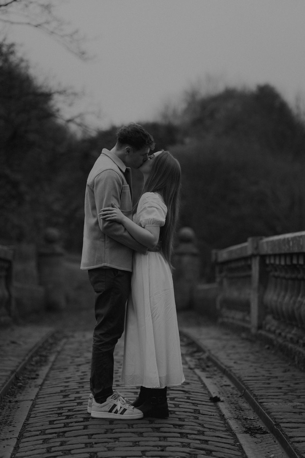Black and white photo of a couple kissing on a cobblestone bridge in a moody outdoor setting.