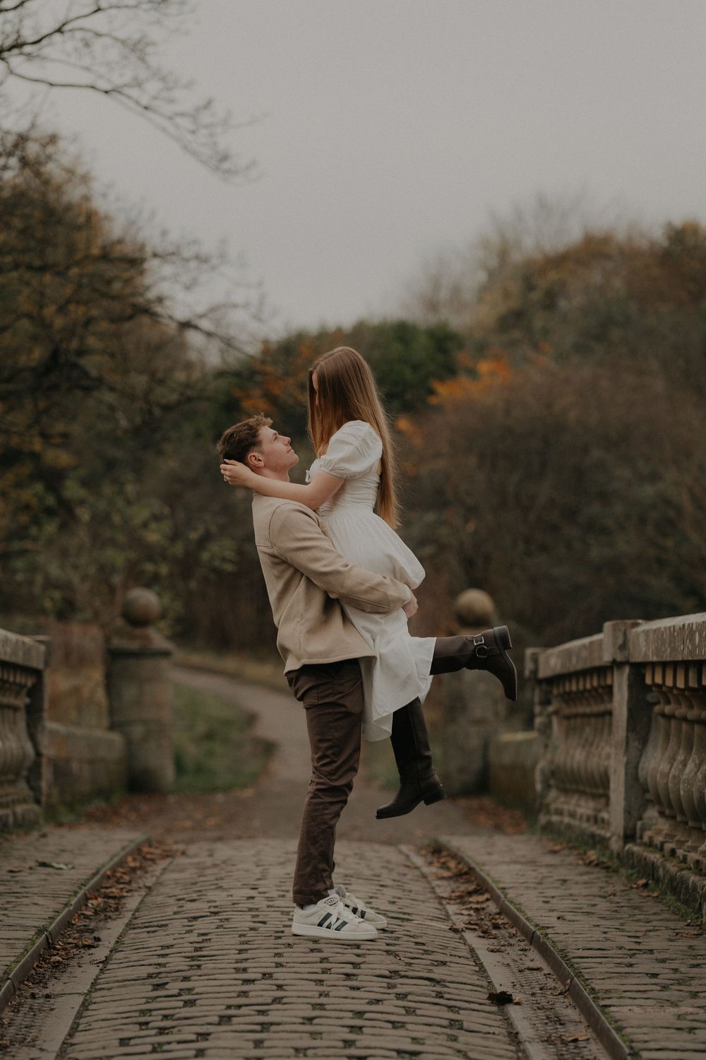 Man lifting woman in white dress on a stone bridge surrounded by autumn trees.
