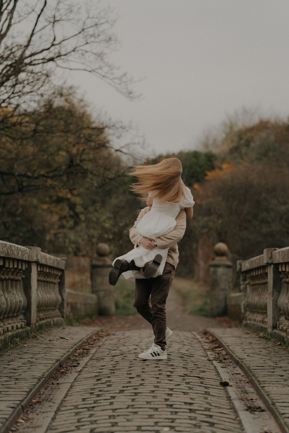 Couple spinning joyfully on a cobblestone bridge in an autumn park setting.