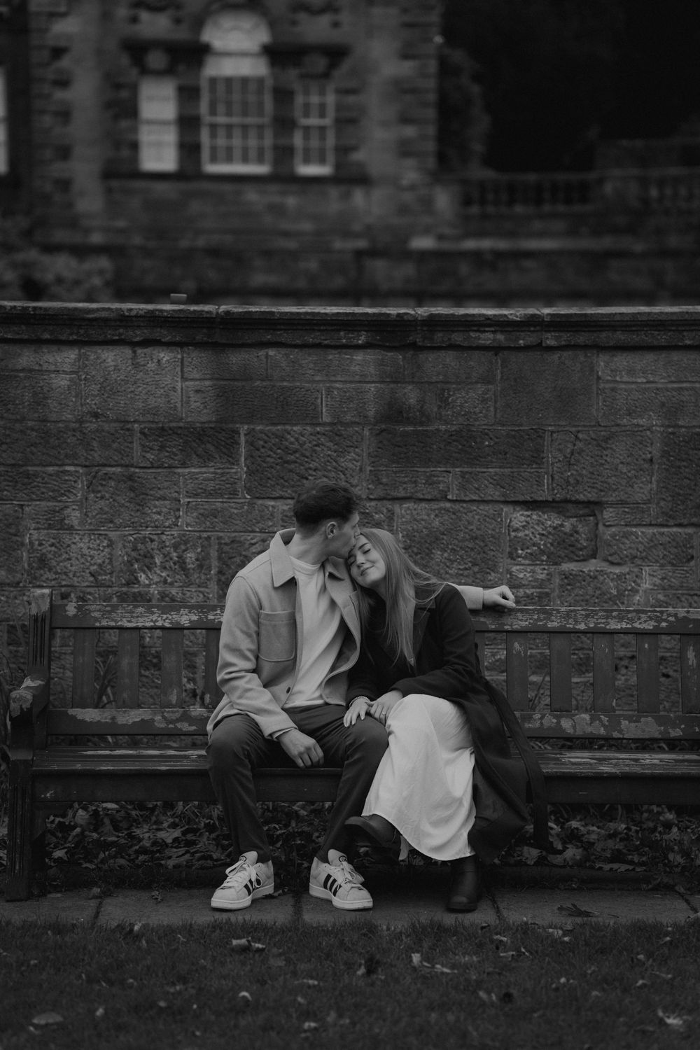 Black and white photo of a couple sitting close on a bench against a stone wall in autumn.