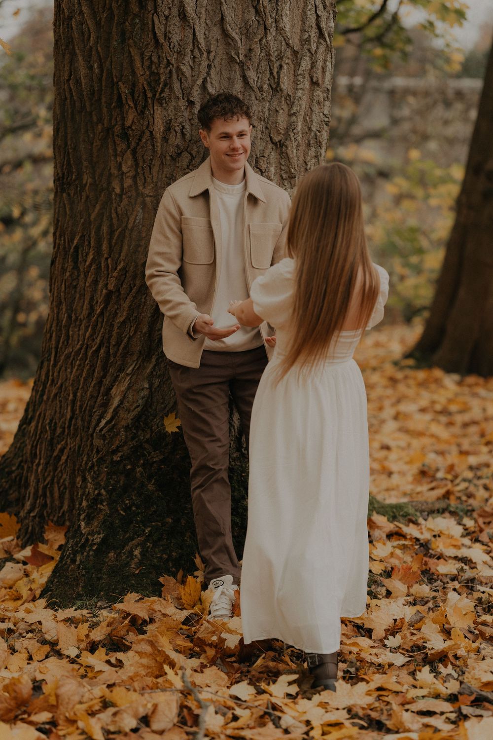 Smiling man leans against a large tree as woman in white dress approaches through fallen leaves.