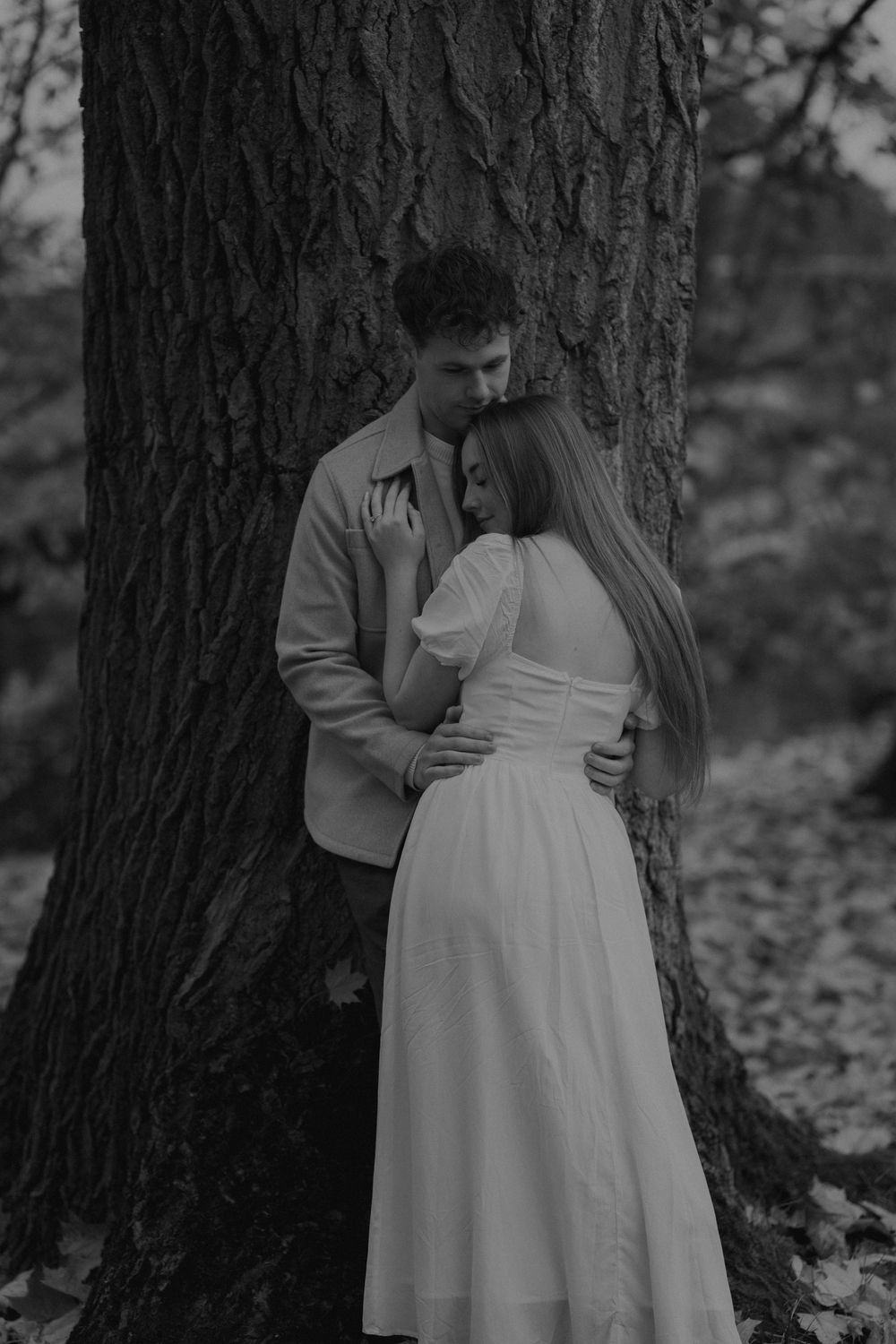 Couple embracing tenderly against large tree trunk in black and white forest portrait.