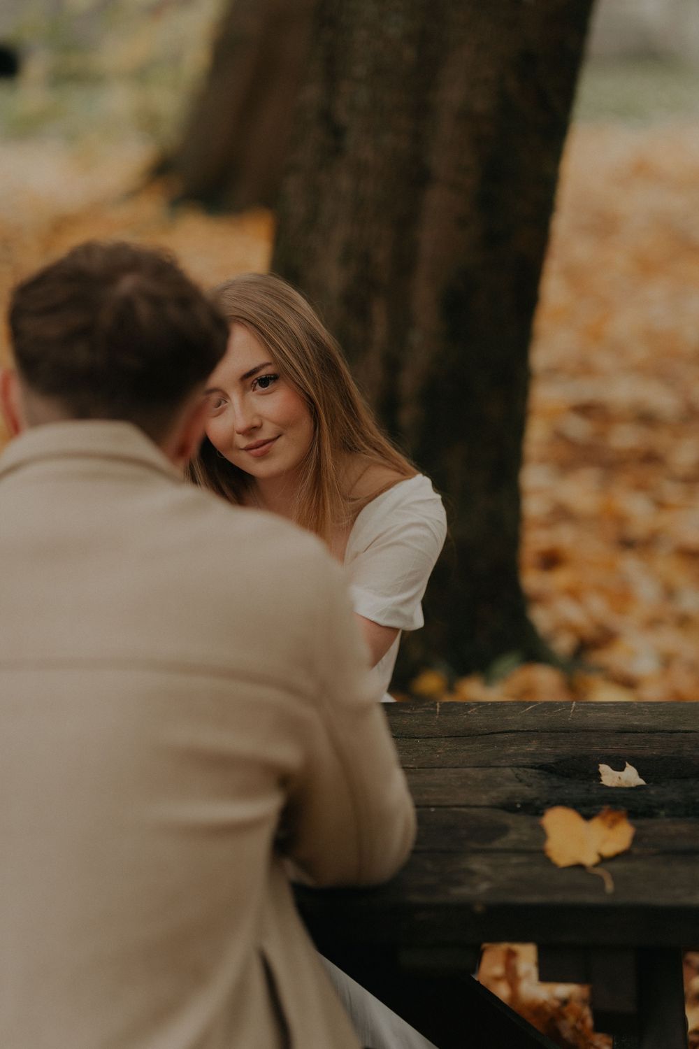 Young woman in a white dress smiling softly at her partner across a wooden bench in an autumn park.