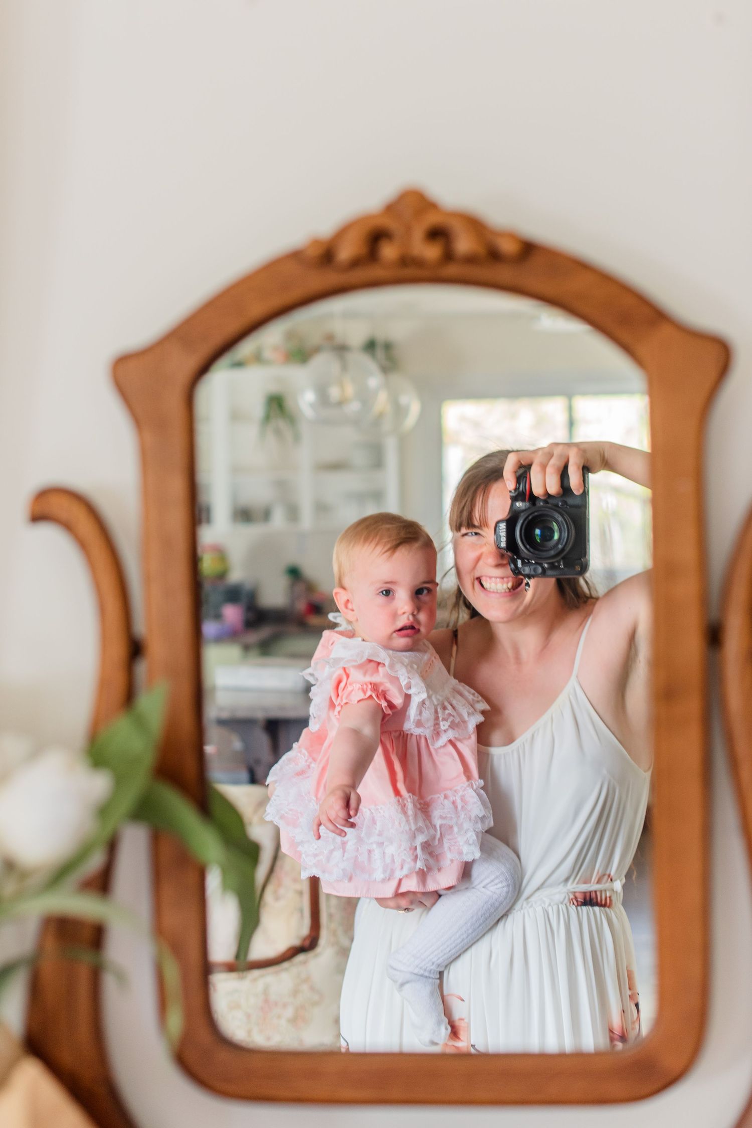 A mother in white dress holds baby in pink ruffle outfit, taking mirror selfie with DSLR camera in ornate wooden frame.