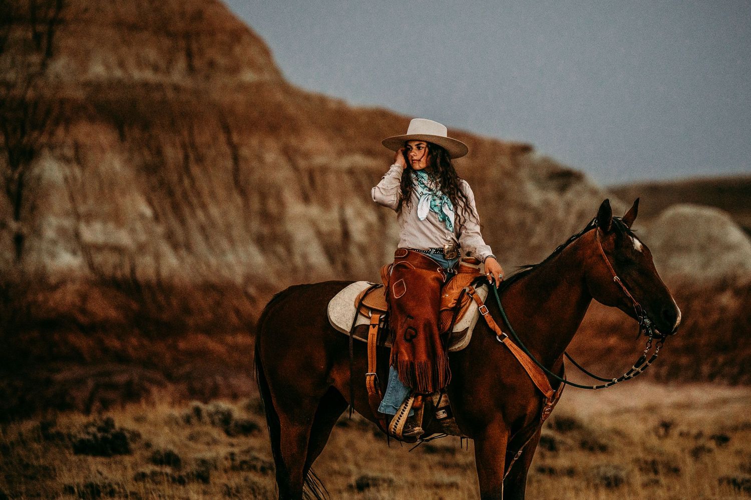 A cowgirl in a white hat rides a brown horse through a rugged desert landscape with rocky badlands in the background.