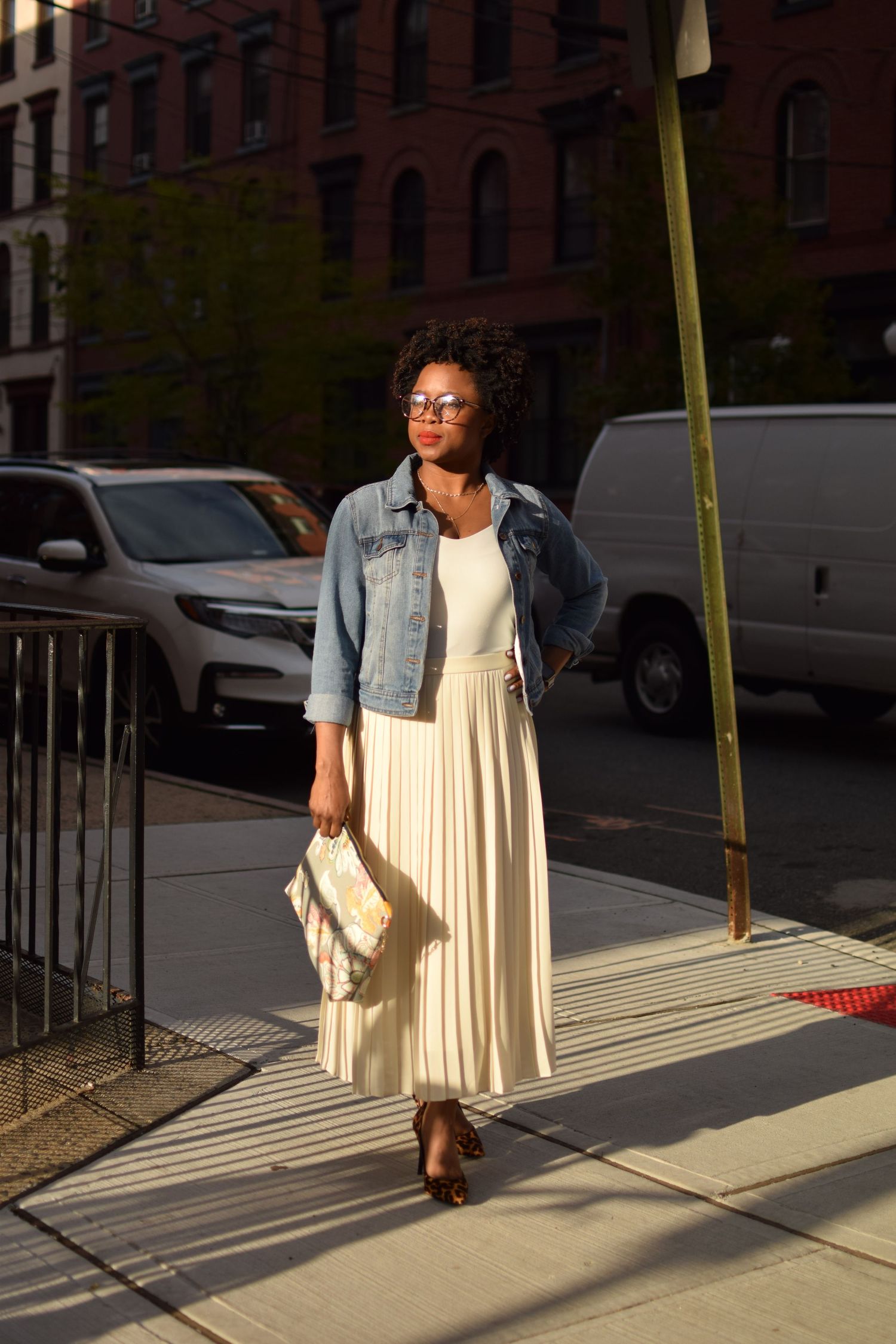 A person wearing a denim jacket and cream pleated midi skirt stands on a sunny sidewalk in an urban setting.