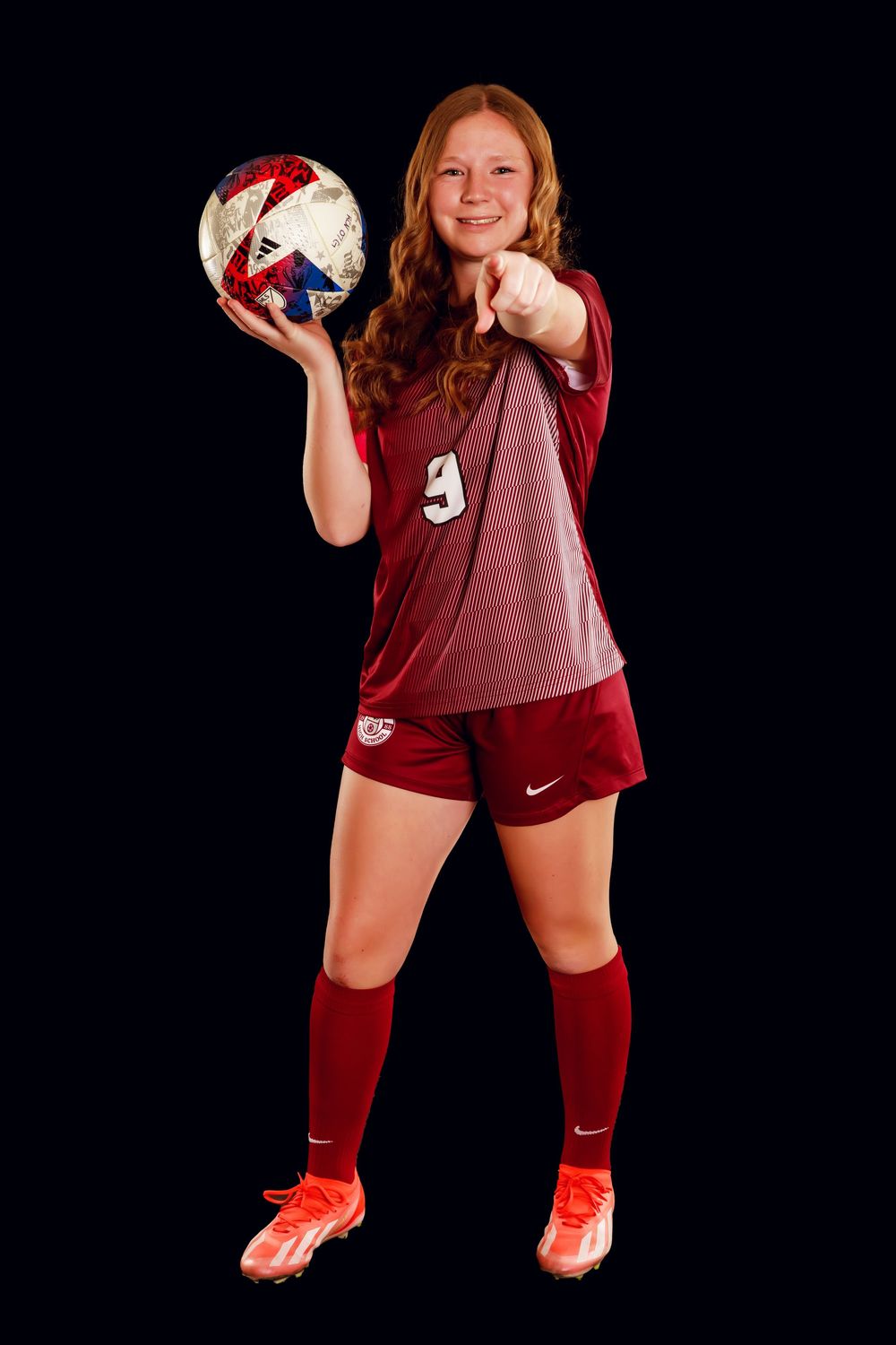 Soccer athlete in maroon uniform poses with soccer ball against dark background.