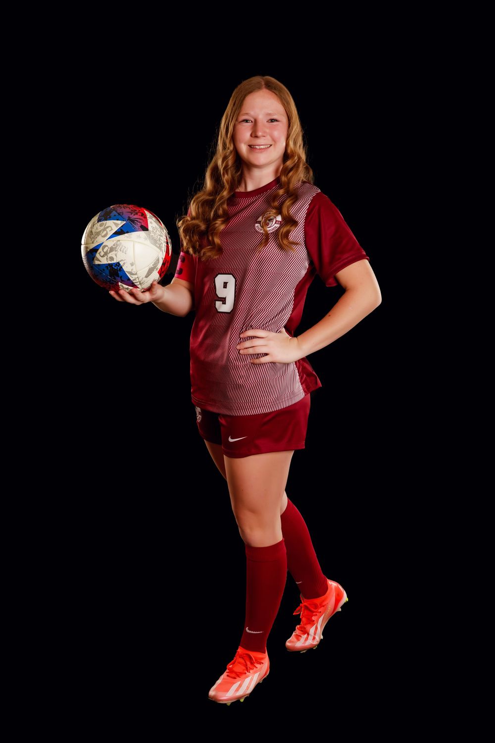 Soccer player in maroon uniform holding ball poses for athletic portrait against black background.