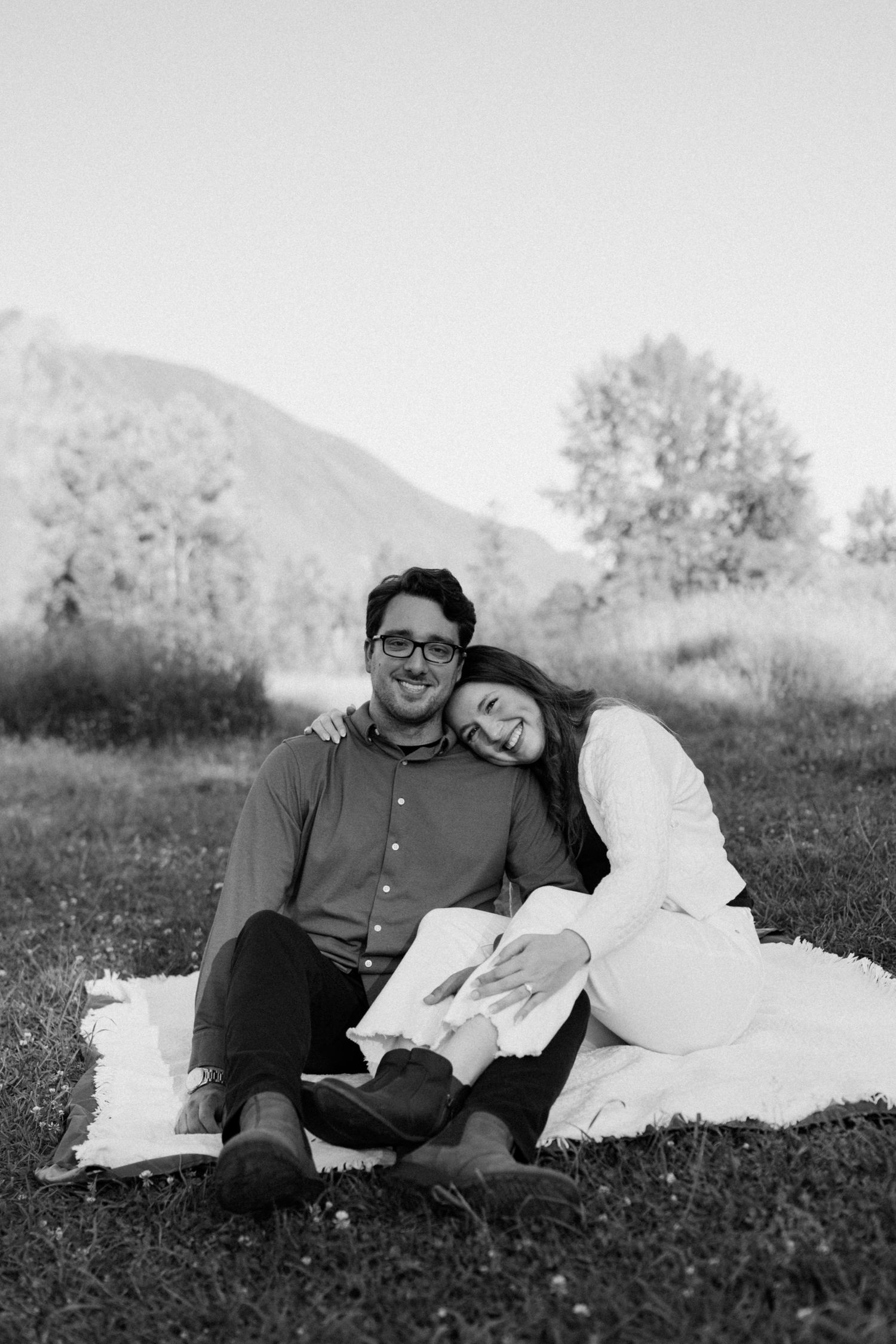 A romantic black and white photo of a couple lounging together on a blanket outdoors with mountains in the background.