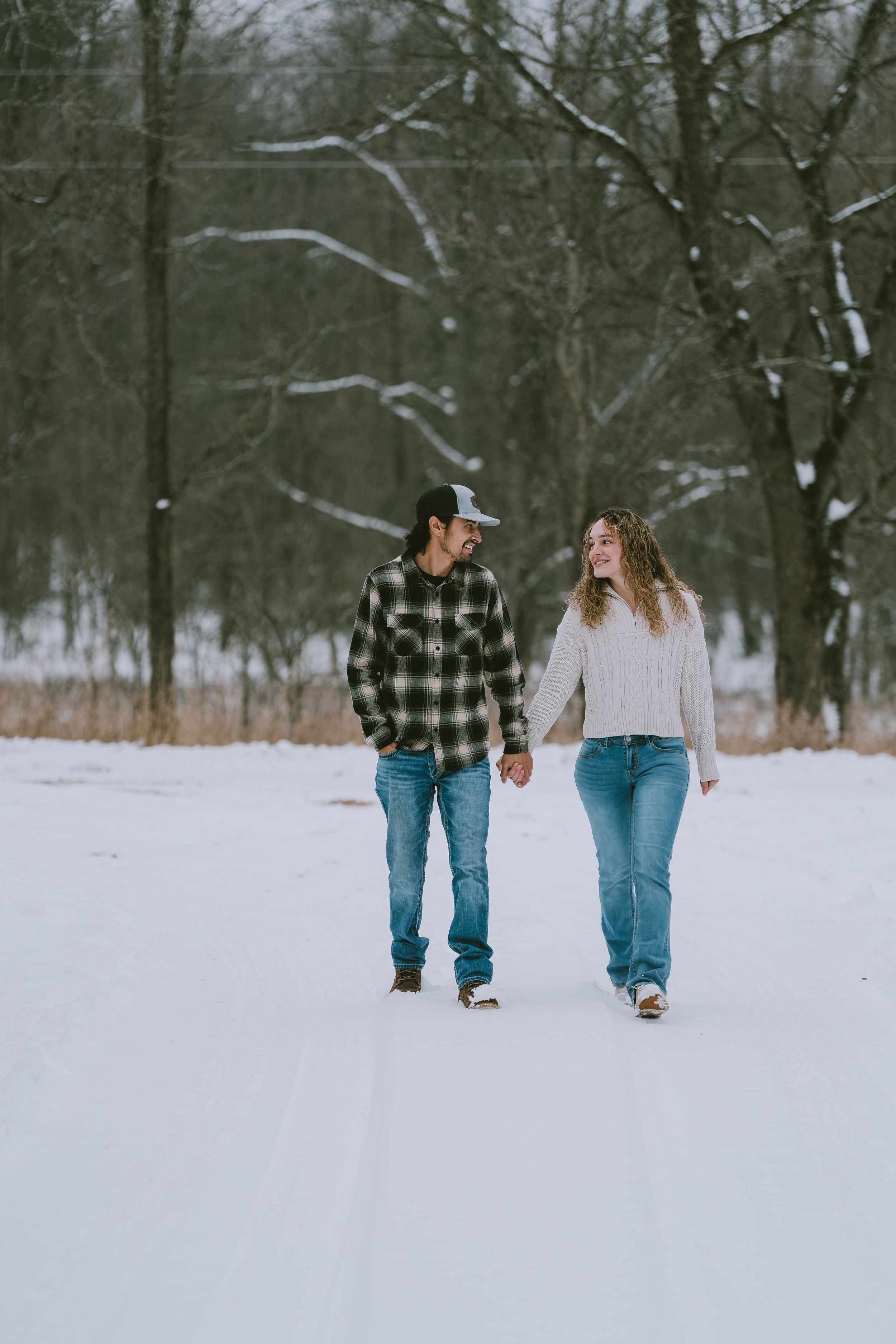 Couple walking hand in hand through a snowy winter forest path wearing casual sweaters and jeans.