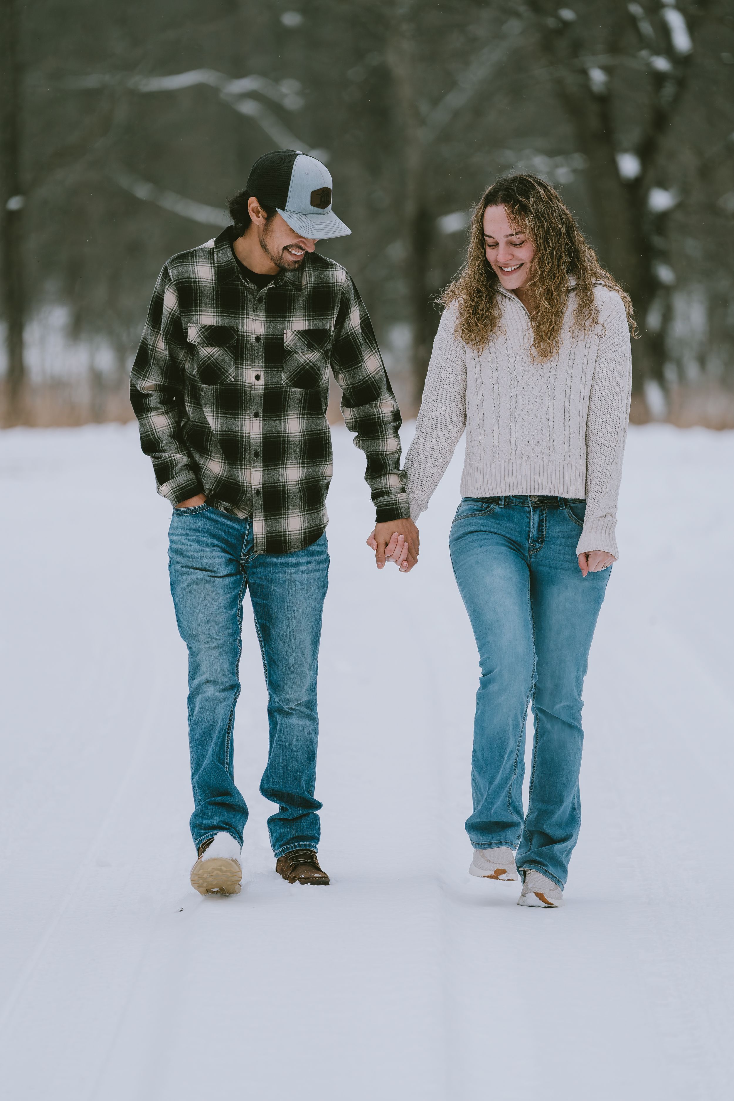 Two people walking together on a snowy path wearing jeans, a plaid flannel and a cream sweater.