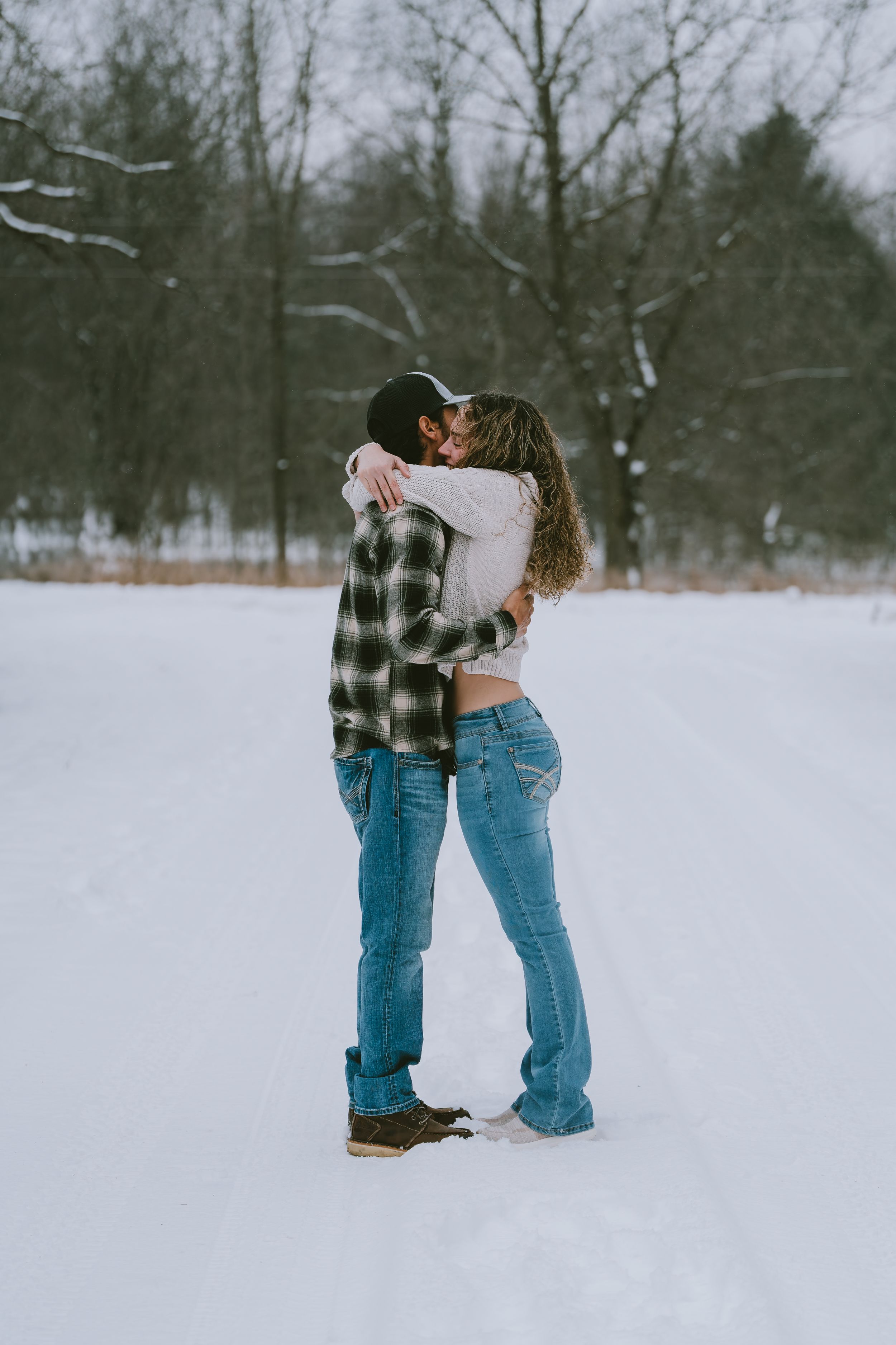 Two people share an intimate embrace while standing in the snow during winter, surrounded by bare trees.