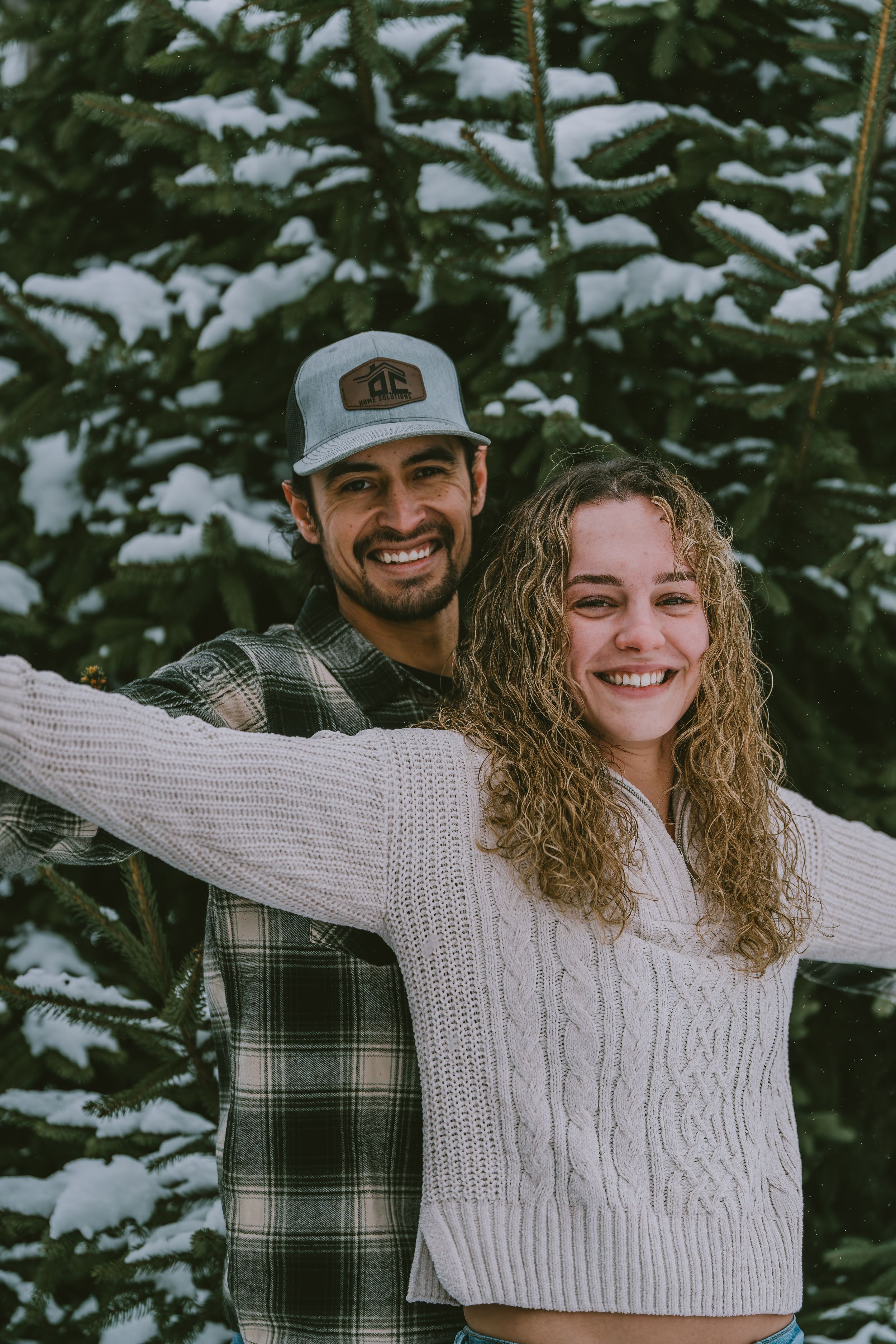 A couple stands together in front of snow-covered evergreen trees, wearing cozy winter sweaters and smiling.