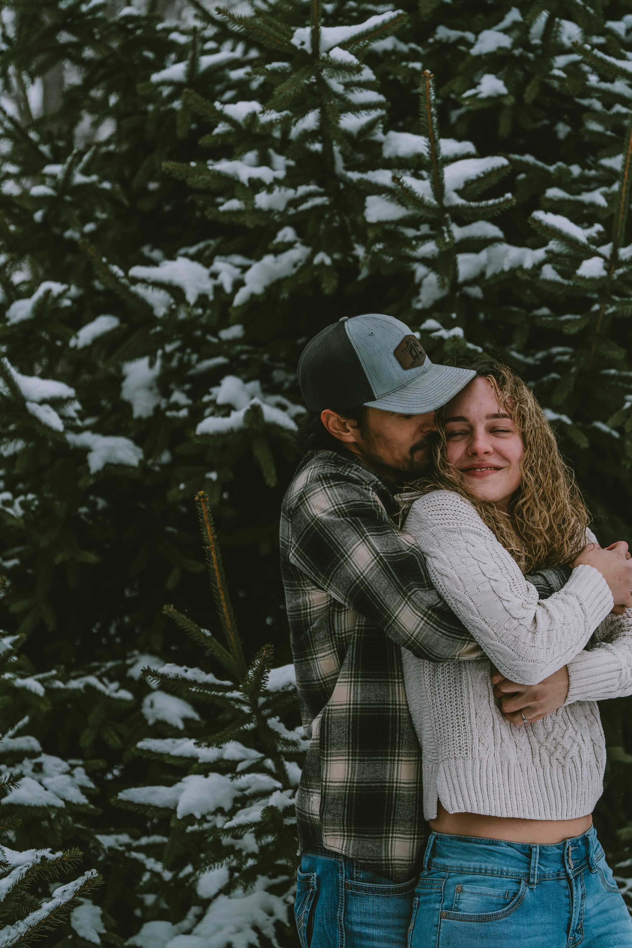 A romantic winter embrace between two people in warm clothing against snow-covered evergreen trees in the background.