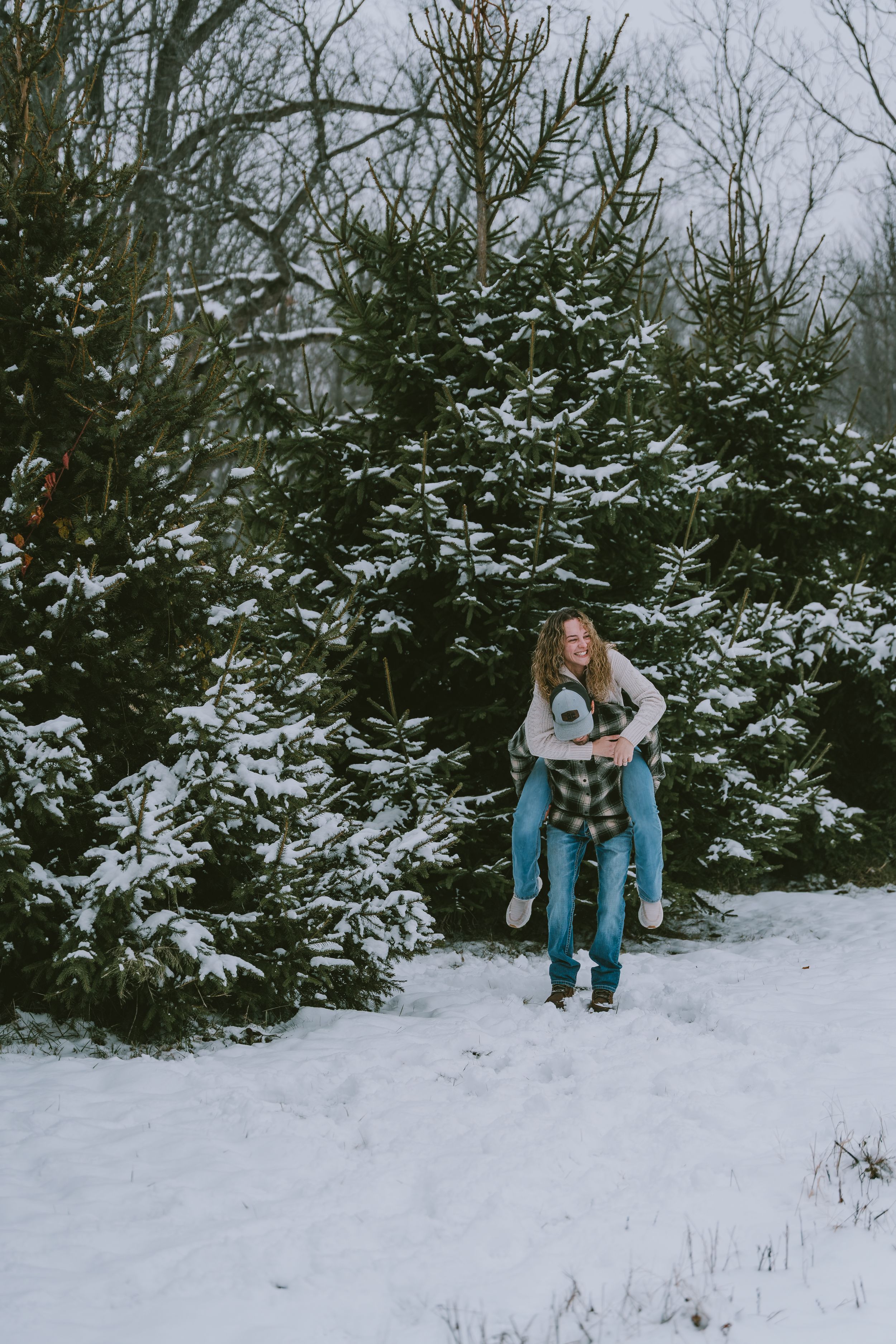 Two people share a piggyback ride in a snowy winter forest setting with snow-covered evergreen trees in the background.